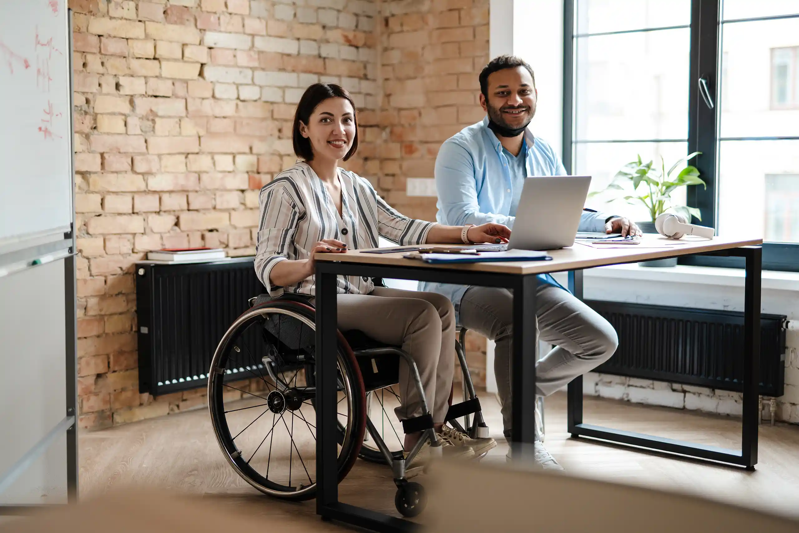 A man and a women smiling to their left, sitting in front of a desk with a laptop. The woman is seated on a wheelchair
