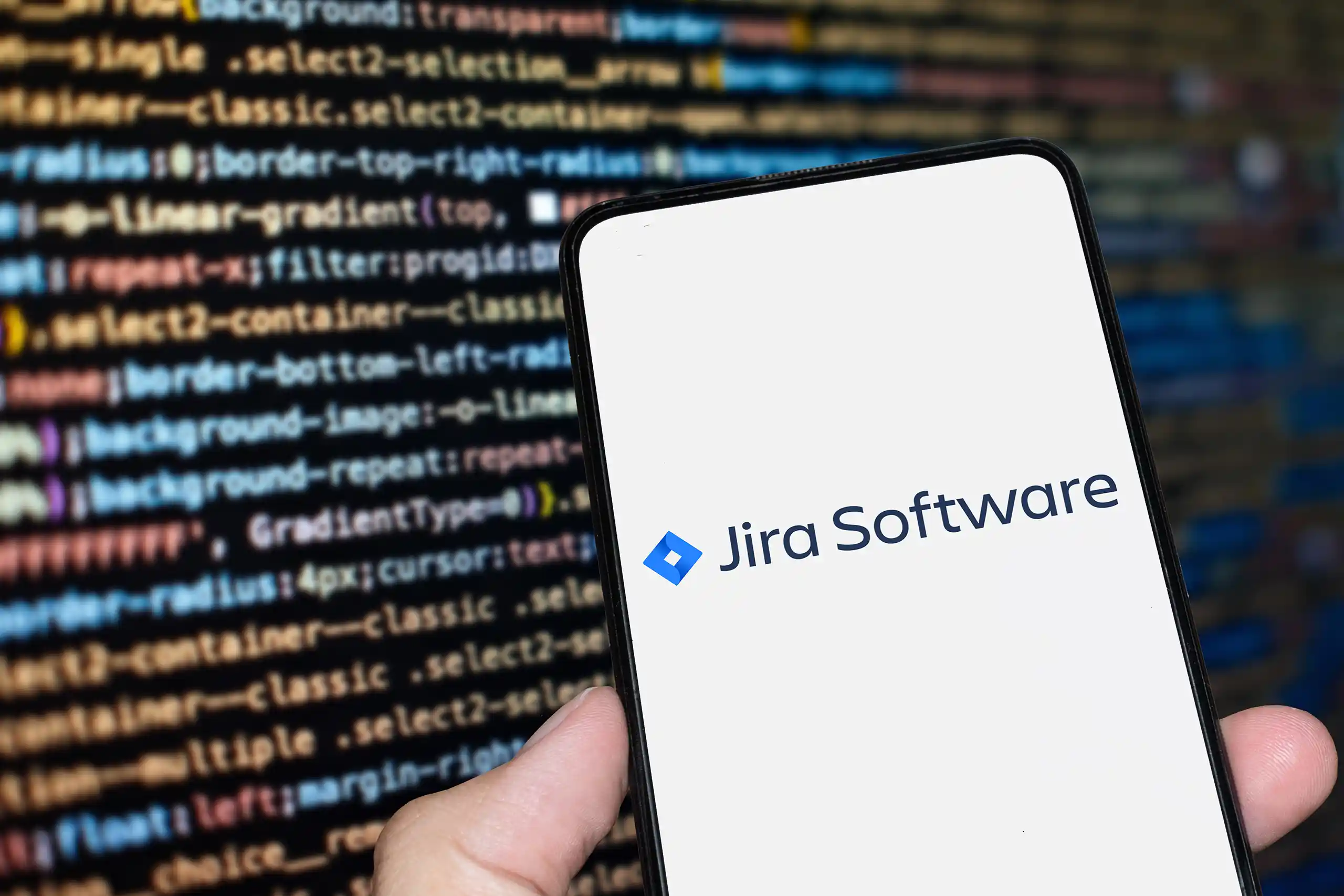 Hand holding a smartphone showing 'Jira Software' with a slightly blurred screen in the background showing CSS style sheet code