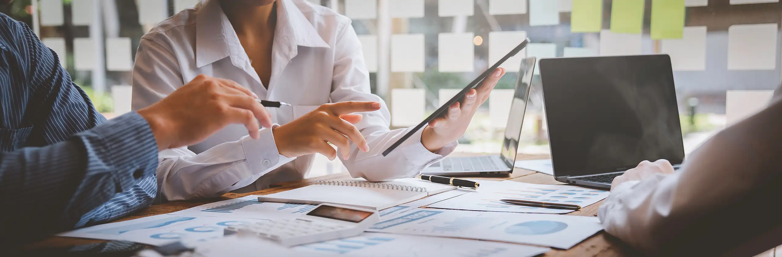 Person on a desk with a laptop and papers pointing to a tablet device next to a person pointing to the device with a pen