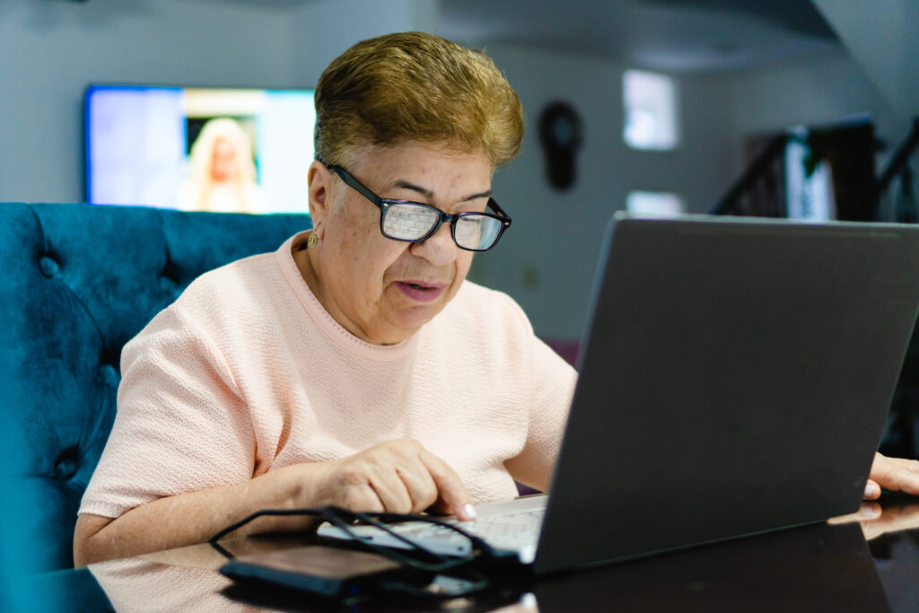 Older woman wearing glasses using a laptop at home, concentrating on the screen with assistive technology nearby.