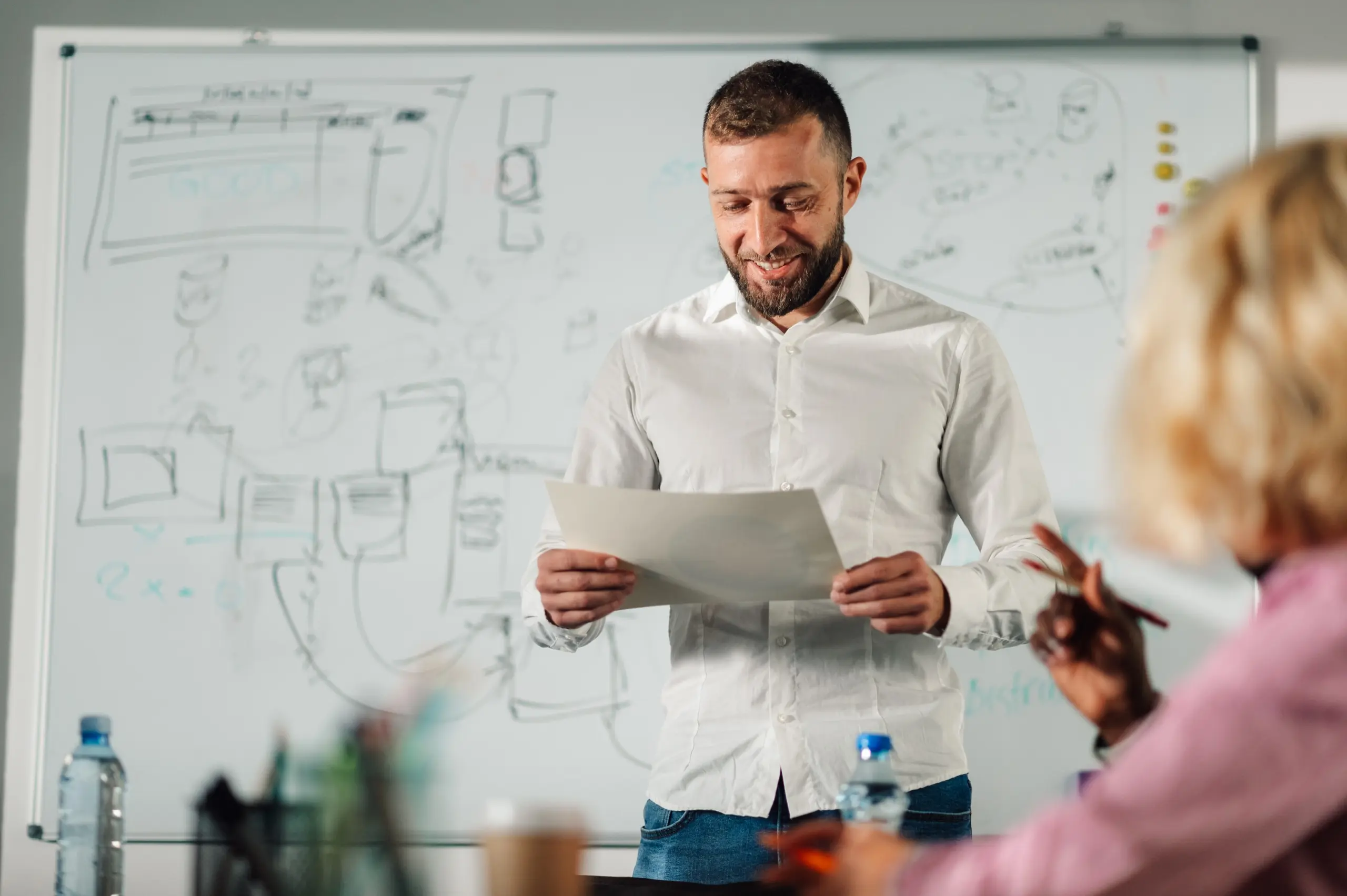 Man with white shirt standing in a room looking down on a sheet of paper he is holding, there is a whiteboard in the background with diagrams and people sitting on a desk in front of the man