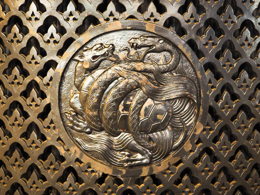 Bronze relief carving of Xuanwu, the mythological Black Tortoise entwined with a snake, set in a patterned background, symbolising mystery and protection.