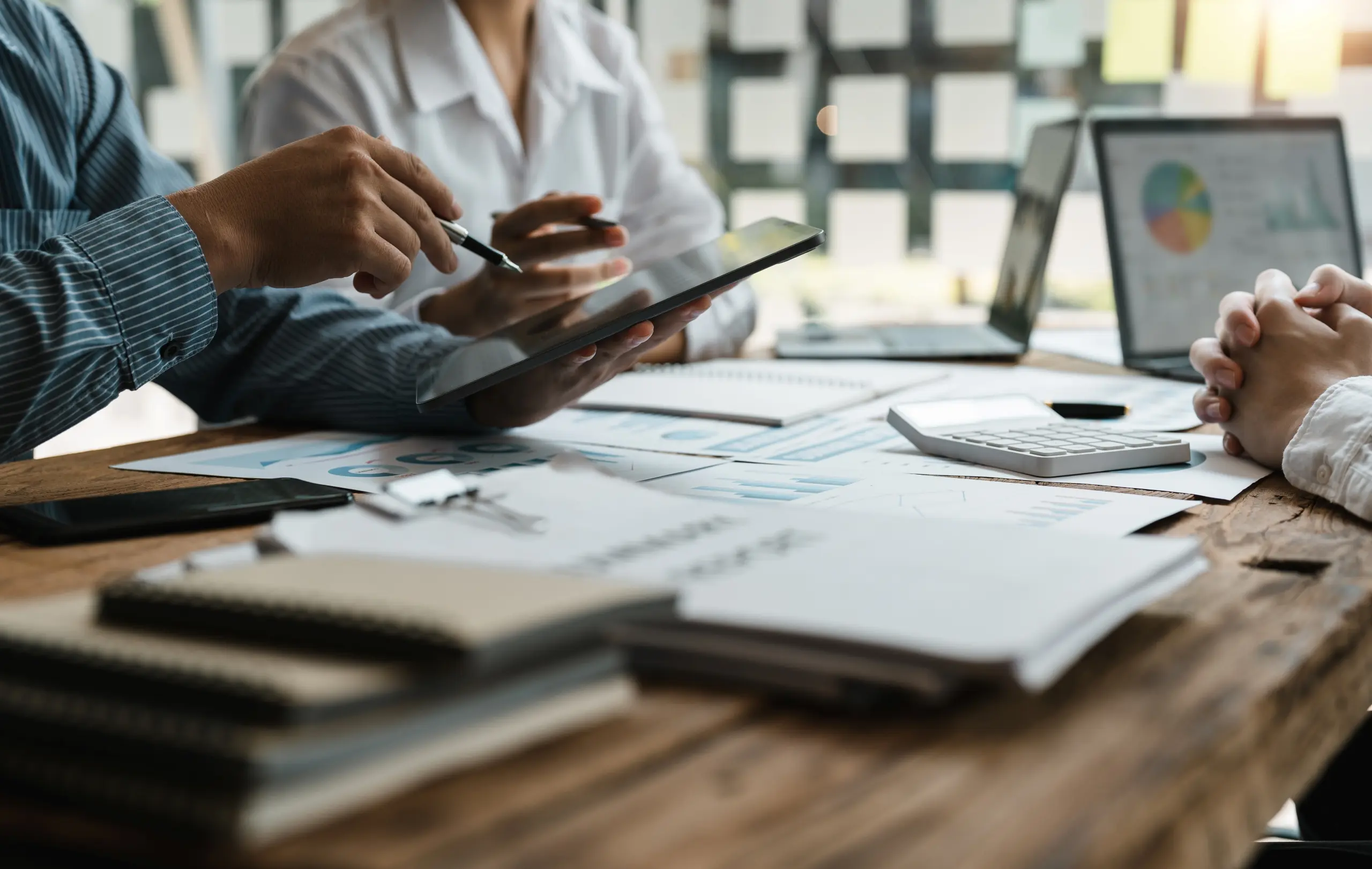 Two people sitting on desk with papers, notebooks, laptops and a calculator, one person holds a tablet device and points to it with a pen together with the person sitting next to them