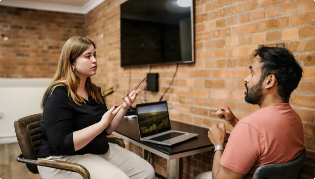 Man and woman seated and signing together; woman is beside a wall desk with an open laptop