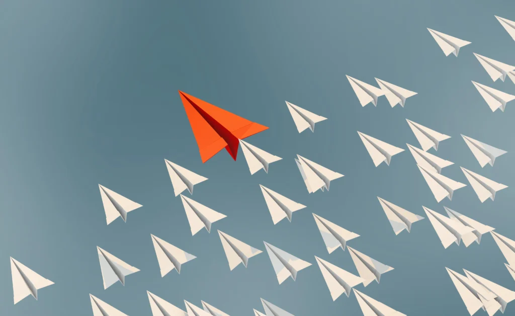 Red paper airplane flying ahead of a group of white paper airplanes.